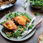 Angled shot of a speckled ceramic plate with two golden-skinned chicken thighs over fresh arugula dressed with agrodolce sauce, on a dark wood table with crusty bread and arugula in the background.