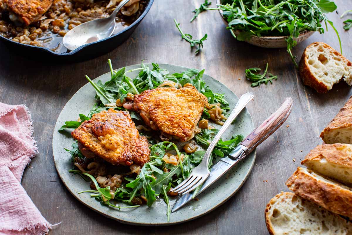 Angled shot of a speckled ceramic plate with two golden-skinned chicken thighs over fresh arugula dressed with agrodolce sauce, on a dark wood table with crusty bread and arugula in the background.