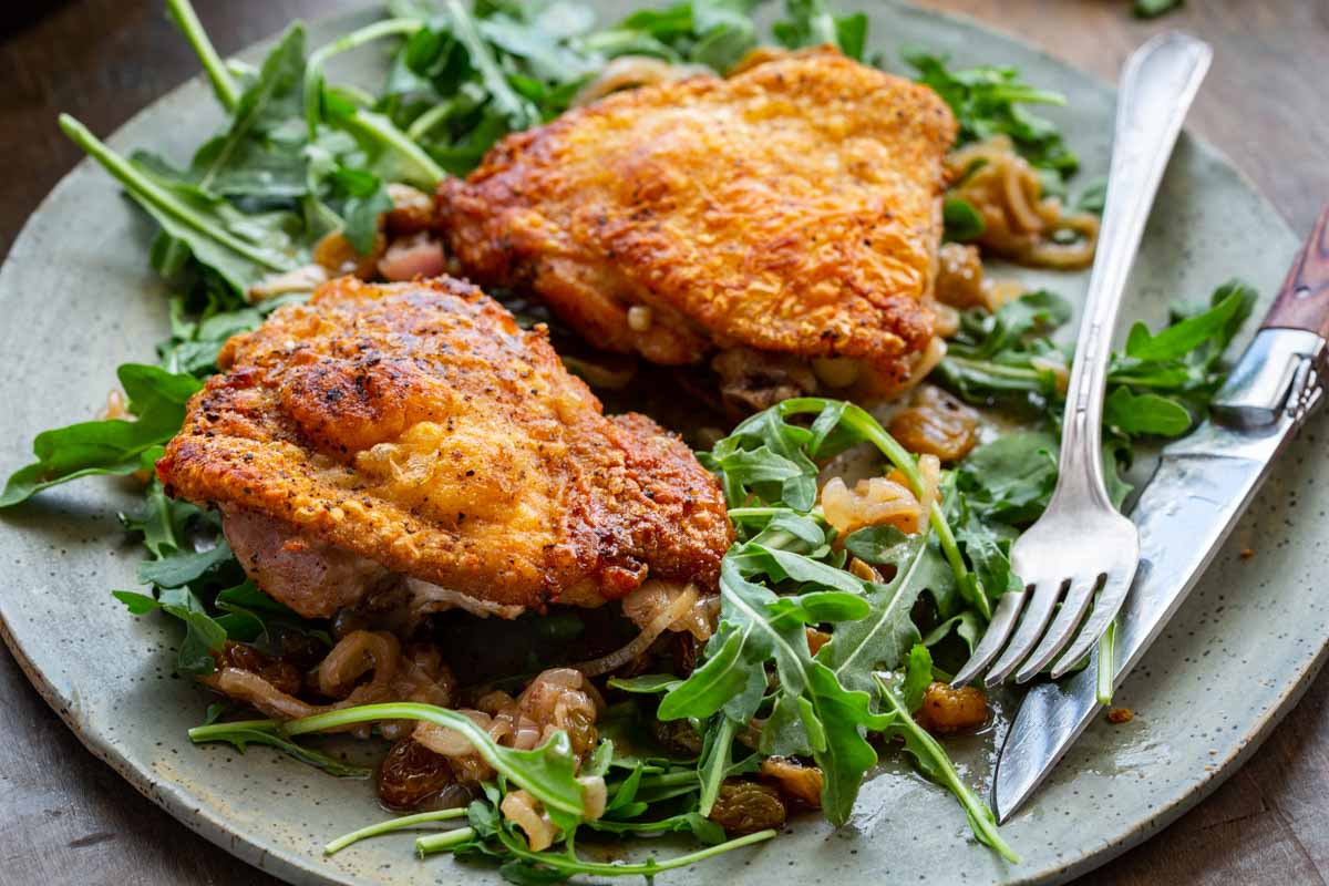Close-up shot of two crispy-skinned chicken agrodolce thighs on a bed of fresh arugula and caramelized shallot-raisin sauce, on a speckled ceramic plate with a fork and knife resting on the side.