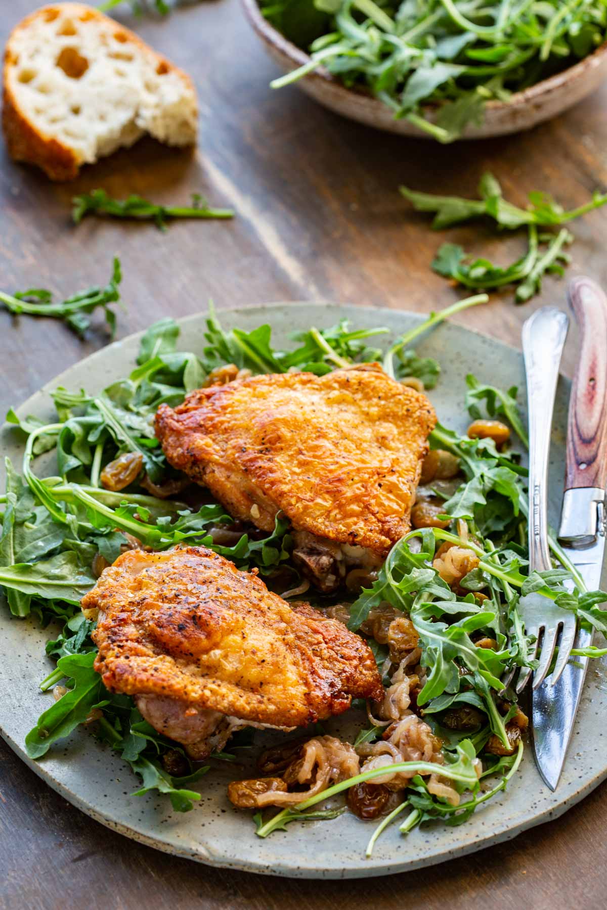 Three-quarter angle shot of two golden-brown chicken thighs served over arugula with agrodolce sauce on a speckled plate, with crusty bread and a bowl of fresh arugula visible in the background on a dark wood surface.