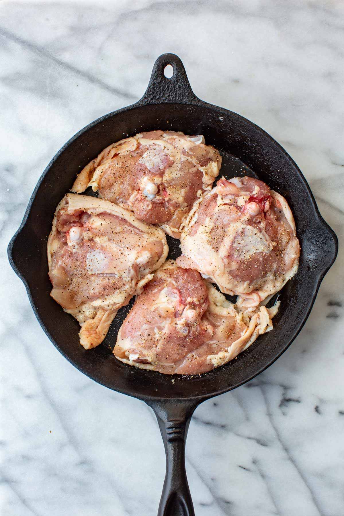 Overhead shot of four raw, bone-in skin-on chicken thighs seasoned with salt and pepper, arranged in a black cast iron skillet on a marble surface.