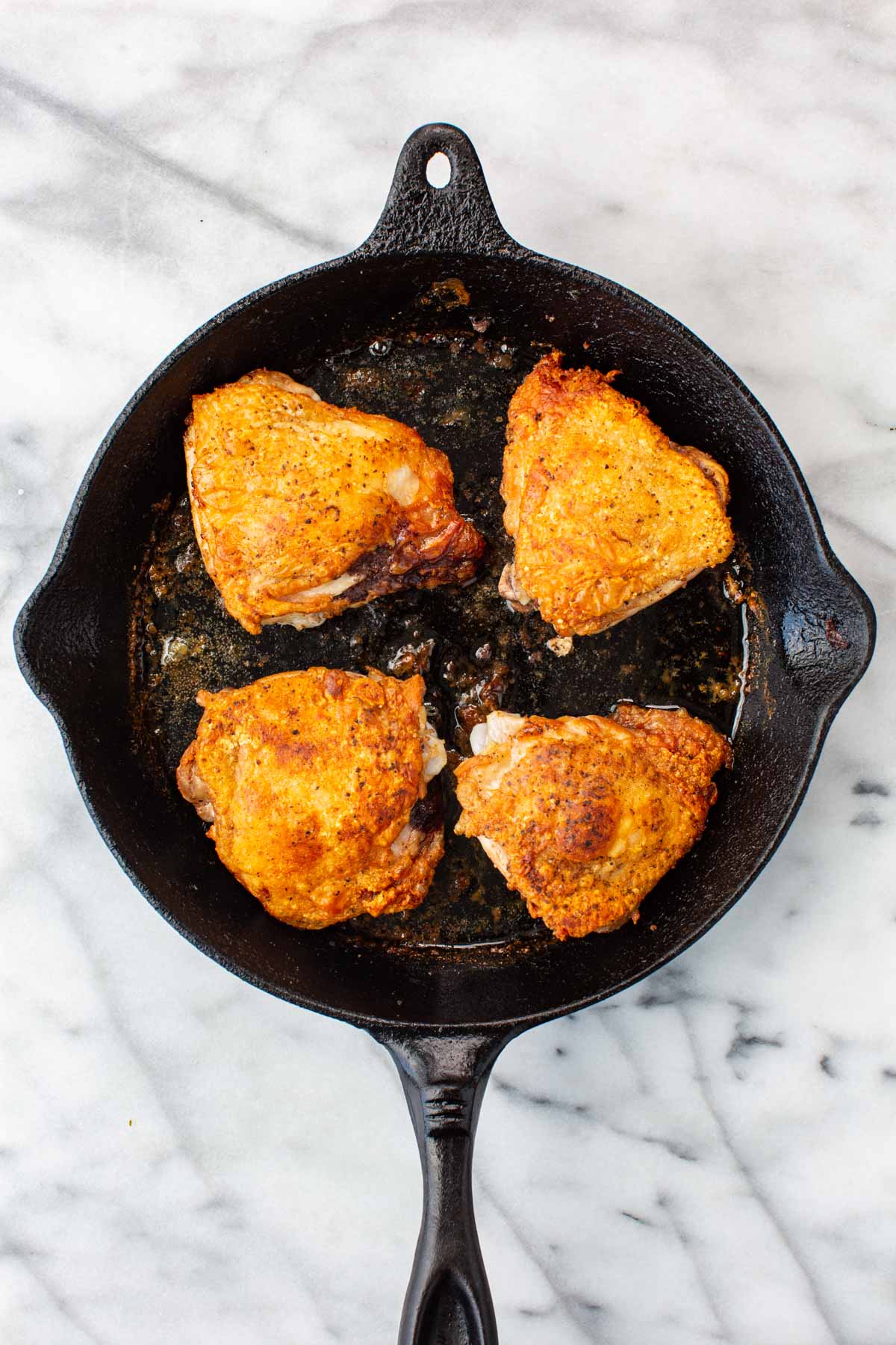Overhead shot of four bone-in, skin-on chicken thighs seared to a deep golden brown, skin side up, in a black cast iron skillet on a marble surface.