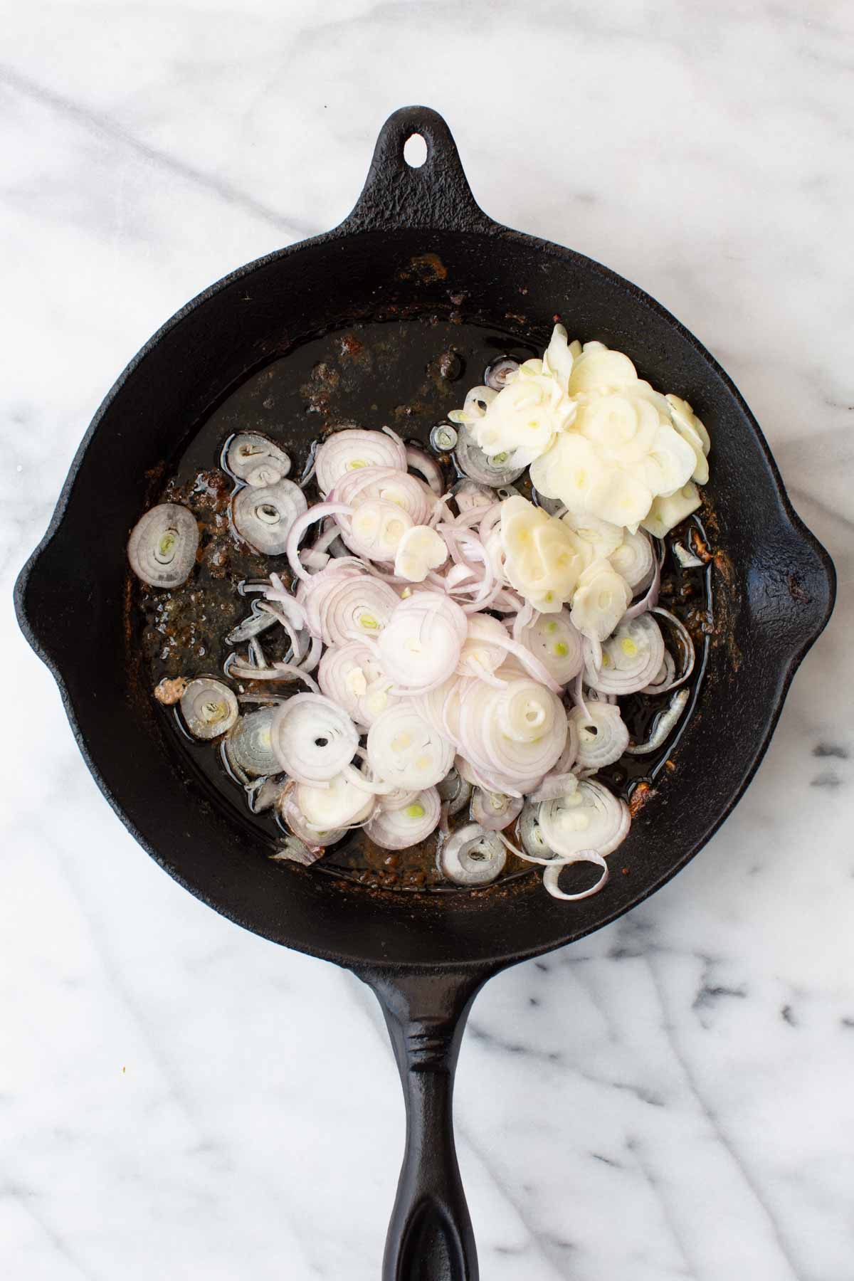 Overhead shot of thinly sliced raw shallots and sliced garlic cloves added to a cast iron skillet with browned drippings on a marble surface.