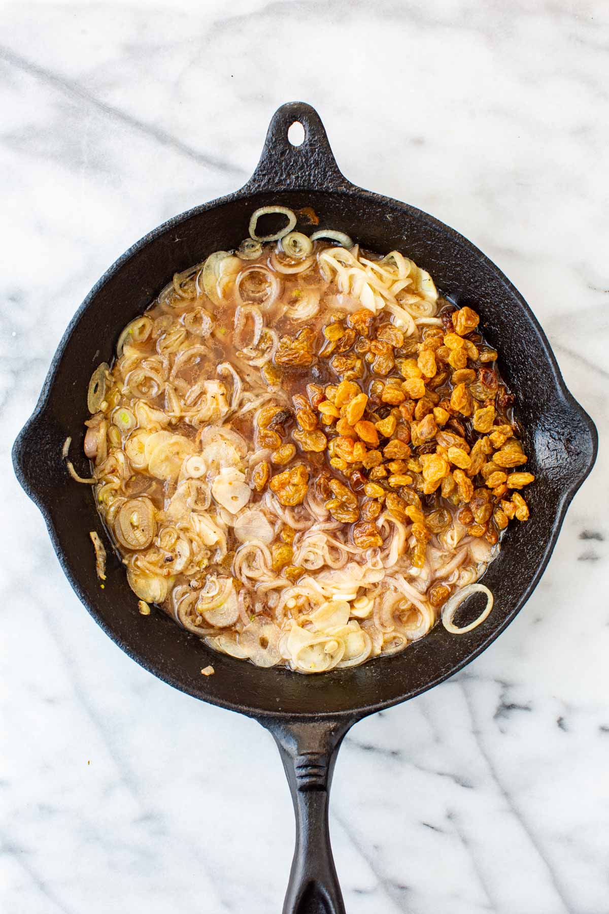 Overhead shot of a cast iron skillet with softened shallots and garlic in a light broth, with golden raisins just added to one side.