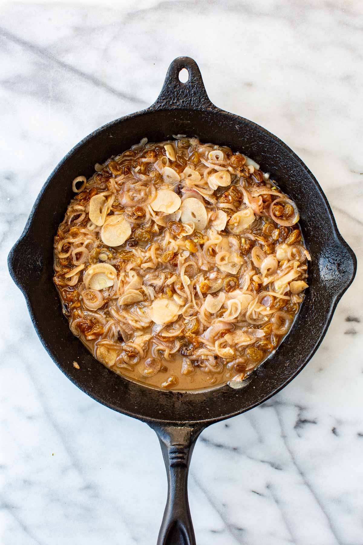 Overhead shot of a cast iron skillet filled with a jammy, golden-brown agrodolce sauce of caramelized shallots, garlic, and plump raisins in reduced braising liquid.