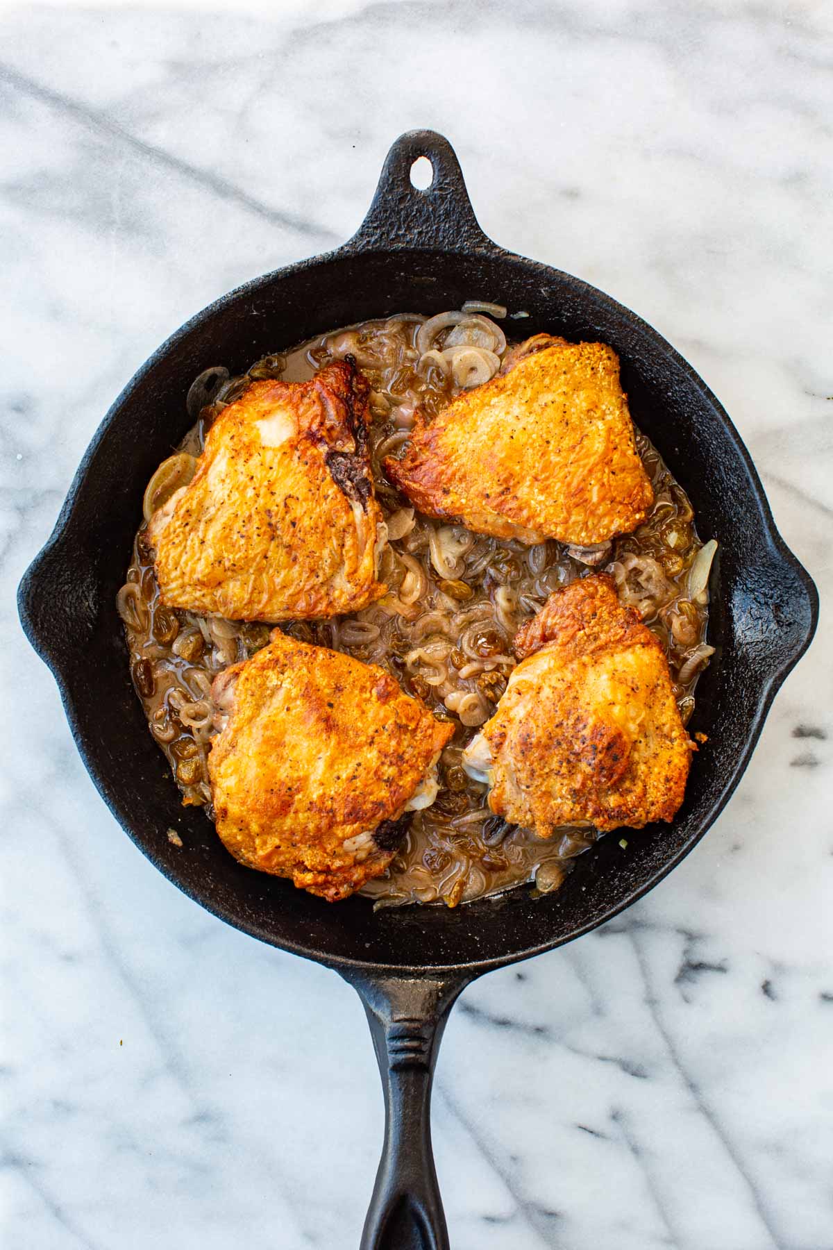 Overhead shot of four seared, golden chicken thighs nestled skin-side up into the agrodolce sauce in a cast iron skillet on a marble surface.