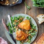 Overhead lifestyle shot of a speckled ceramic plate with two chicken agrodolce thighs served over fresh arugula and spooned with the shallot-raisin pan sauce, set on a rustic dark wood table alongside crusty bread, a bowl of arugula, and the cast iron skillet in the background.