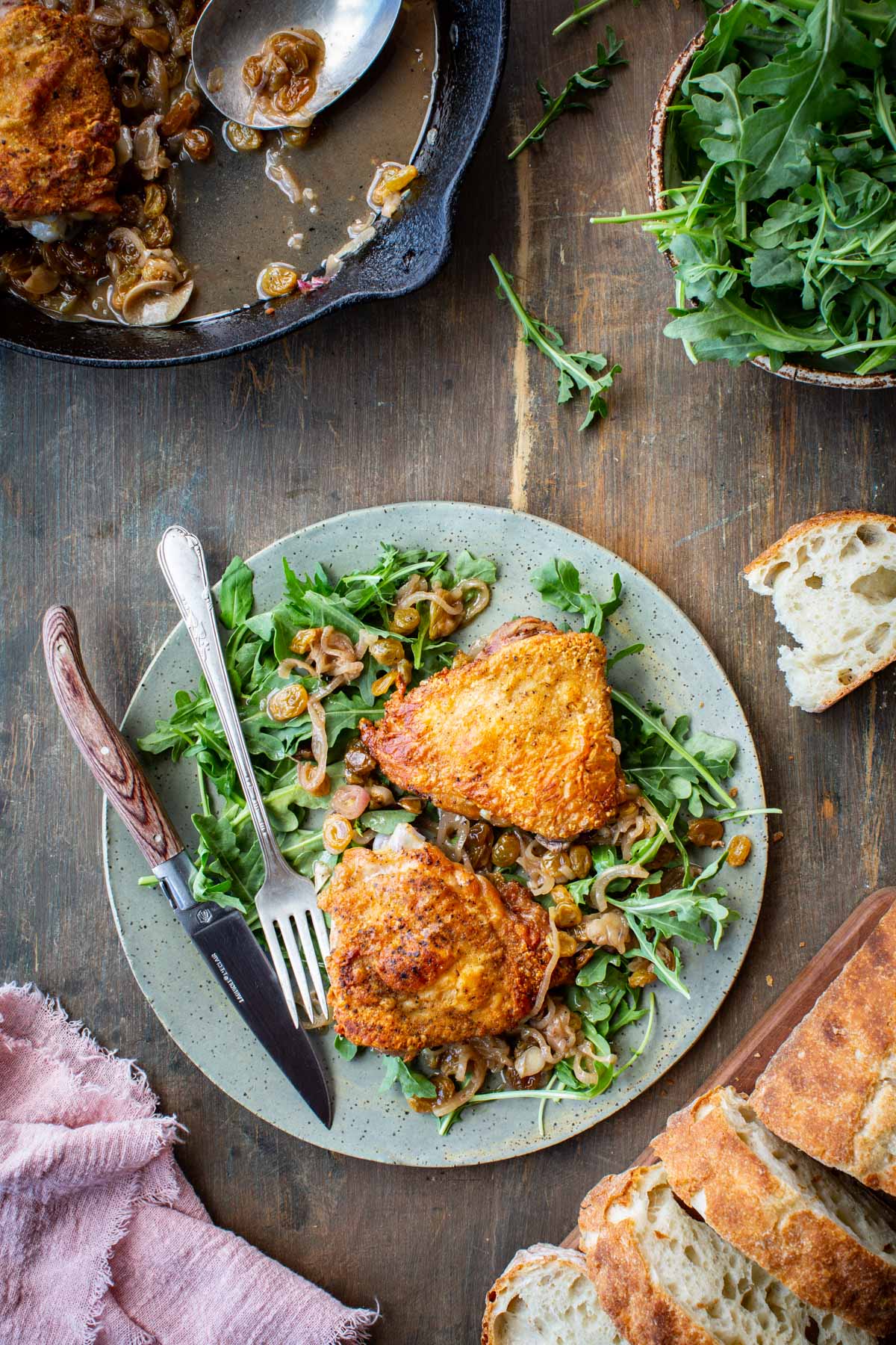 Overhead lifestyle shot of a speckled ceramic plate with two chicken agrodolce thighs served over fresh arugula and spooned with the shallot-raisin pan sauce, set on a rustic dark wood table alongside crusty bread, a bowl of arugula, and the cast iron skillet in the background.