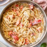 Overhead view of a large stainless steel skillet filled with creamy lobster linguine, garnished with fresh herbs. Chunks of bright red-orange lobster are nestled throughout the pasta. A block of Parmigiano and a box grater sit in the lower left corner, with fresh basil sprigs to the right and a pink linen napkin in the upper right.
