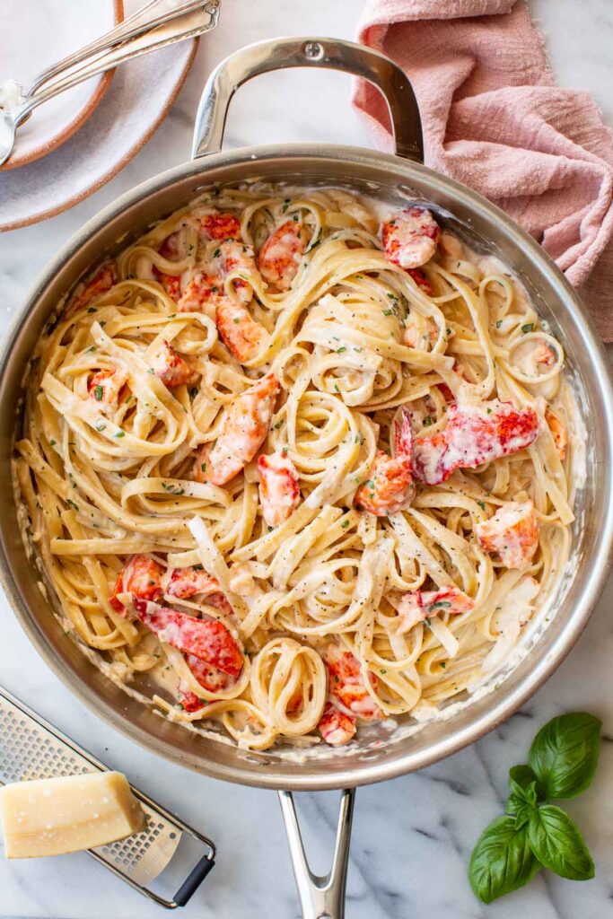 Overhead view of a large stainless steel skillet filled with creamy lobster linguine, garnished with fresh herbs. Chunks of bright red-orange lobster are nestled throughout the pasta. A block of Parmigiano and a box grater sit in the lower left corner, with fresh basil sprigs to the right and a pink linen napkin in the upper right.
