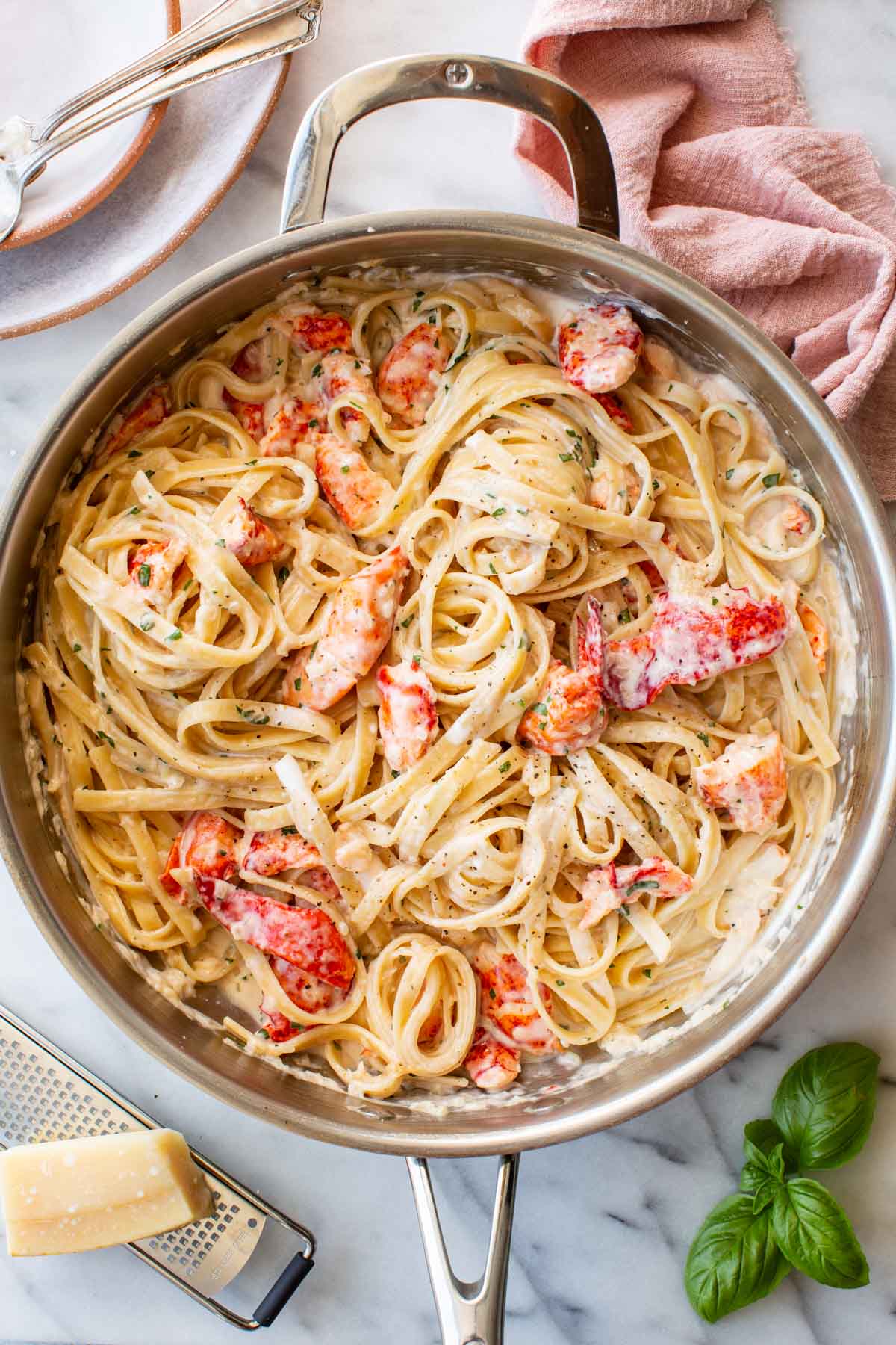 Overhead view of a large stainless steel skillet filled with creamy lobster linguine, garnished with fresh herbs. Chunks of bright red-orange lobster are nestled throughout the pasta. A block of Parmigiano and a box grater sit in the lower left corner, with fresh basil sprigs to the right and a pink linen napkin in the upper right.