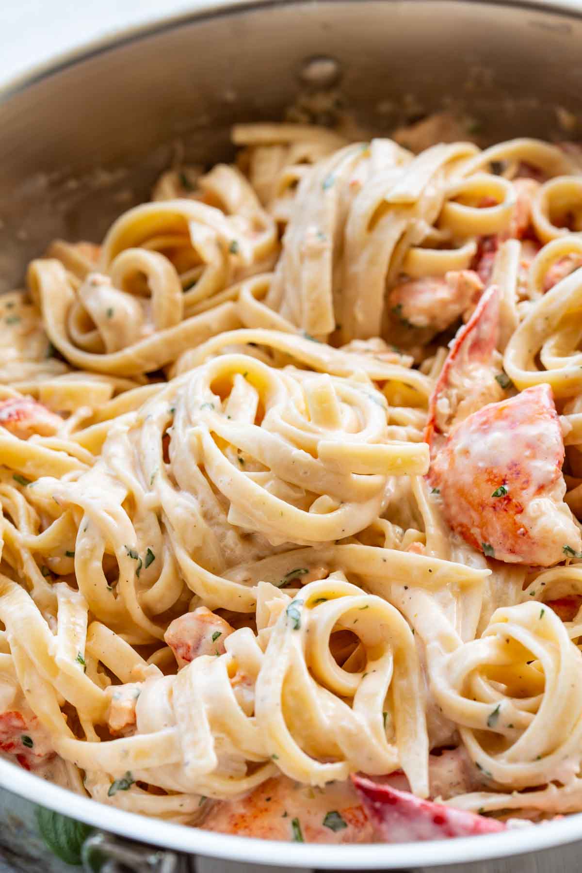 Close-up of creamy lobster pasta in a pan, showing silky linguine coated in a rich white sauce with visible herbs. Several large pieces of cooked lobster claw and tail meat are tucked among the noodles.