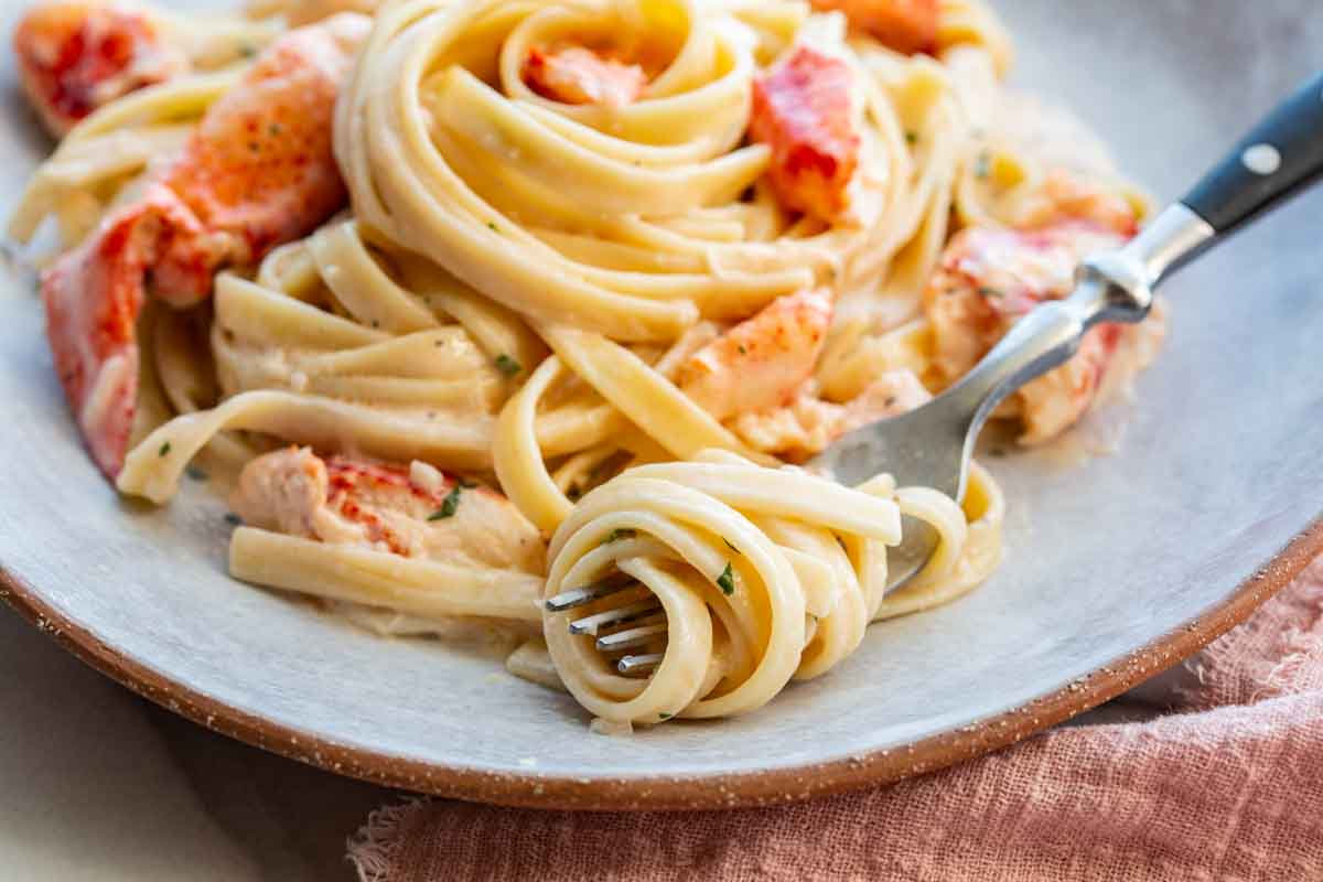 A fork twirled with creamy lobster linguine rests on a rustic ceramic plate. The foreground shows the fork up close with noodles wound around the tines, while additional lobster pieces and more pasta are visible in the background.