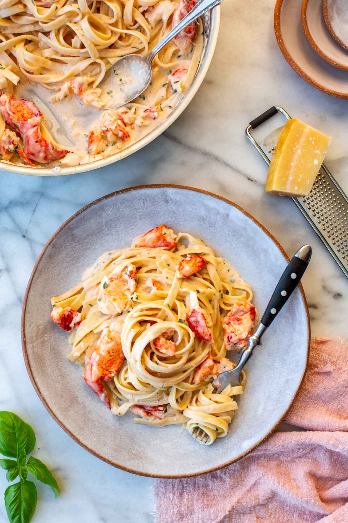 Overhead flat lay of a plated serving of lobster pasta on a gray ceramic plate alongside the larger skillet in the upper left corner. A box grater and wedge of Parmigiano sit to the right, with fresh basil leaves in the lower left and a pink linen napkin to the right.