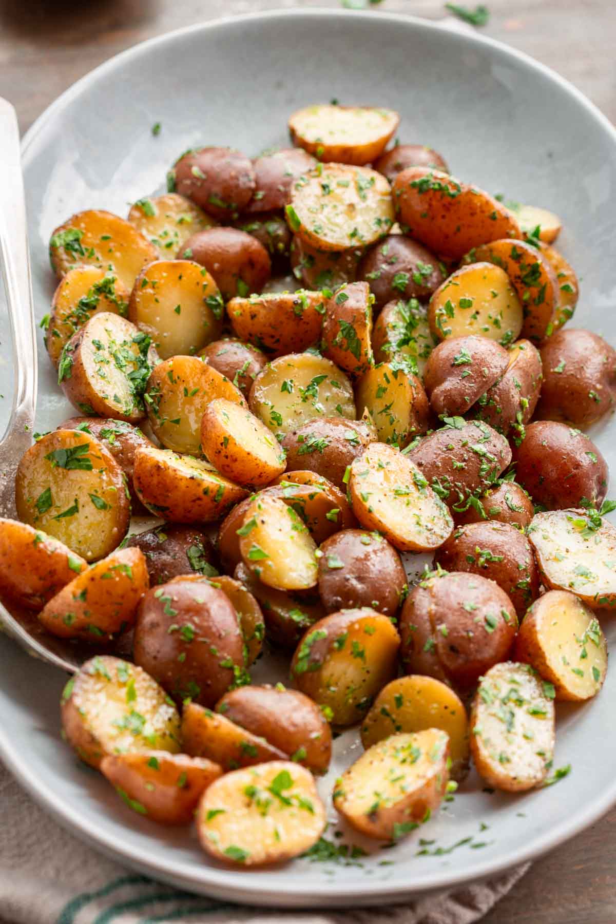 Angled view of buttered parsley potatoes in a light gray bowl, showing their golden cut sides and herb-flecked coating, with a silver serving spoon resting on the side.