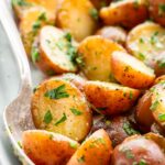Extreme close-up of a silver spoon scooping halved baby potatoes coated in butter and parsley from a serving dish, highlighting the glossy, herb-covered cut surfaces.