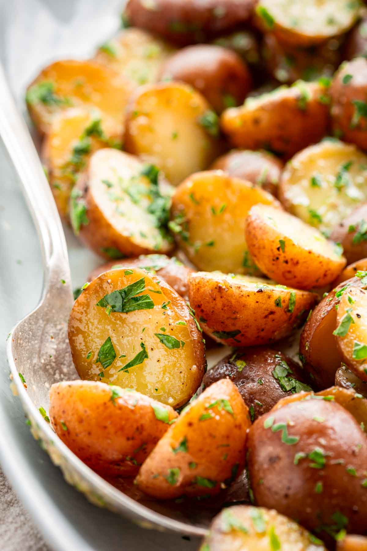 Extreme close-up of a silver spoon scooping halved baby potatoes coated in butter and parsley from a serving dish, highlighting the glossy, herb-covered cut surfaces.