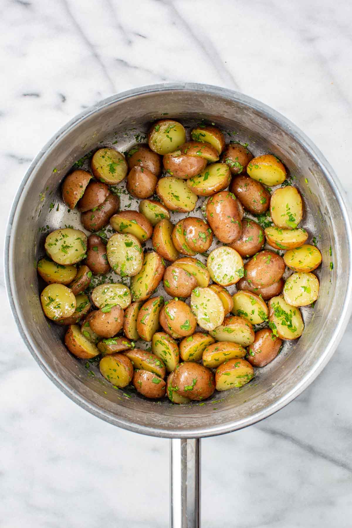 Halved baby red potatoes tossed with melted butter and freshly chopped parsley in a stainless steel saucepan, viewed from above on a white marble surface.