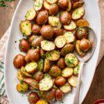 Overhead view of buttered parsley potatoes served on a white oval platter with a vintage silver spoon, surrounded by a small wooden bowl of flaky salt and a bowl of chopped parsley on a wooden surface.