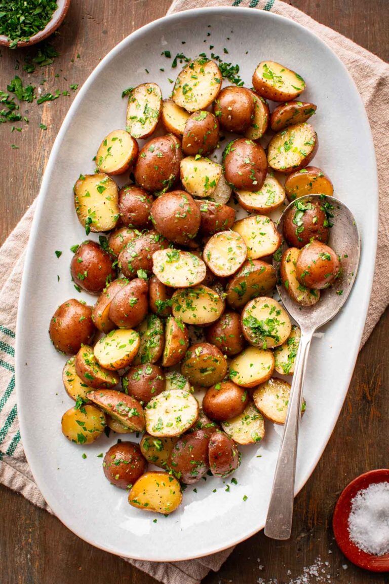 Overhead view of buttered parsley potatoes served on a white oval platter with a vintage silver spoon, surrounded by a small wooden bowl of flaky salt and a bowl of chopped parsley on a wooden surface.