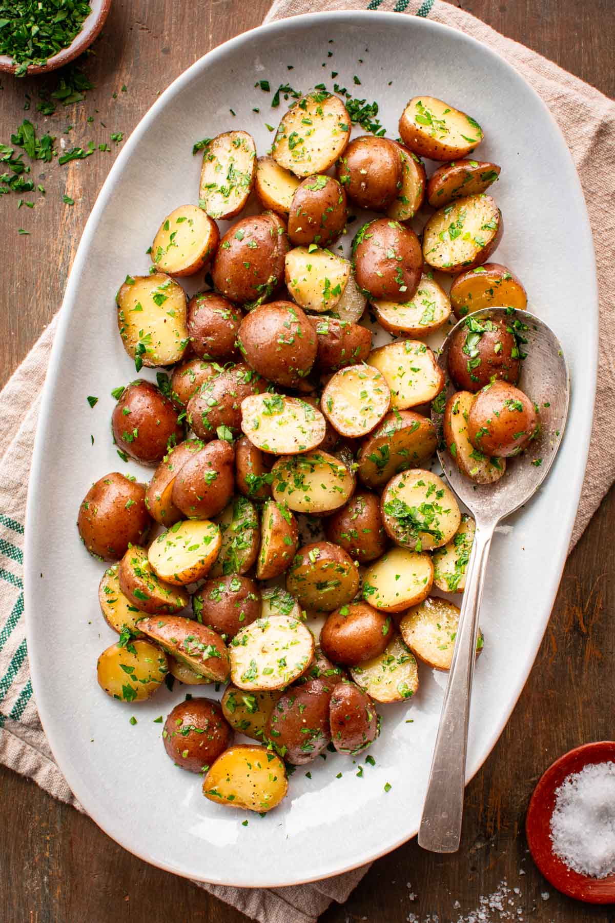 Overhead view of buttered parsley potatoes served on a white oval platter with a vintage silver spoon, surrounded by a small wooden bowl of flaky salt and a bowl of chopped parsley on a wooden surface.