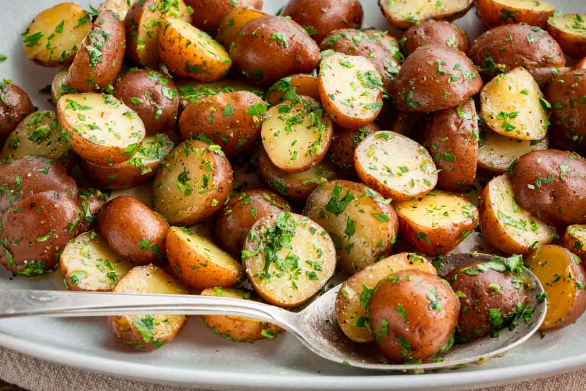 Close-up of halved baby red potatoes generously coated in butter and chopped fresh parsley, piled on a white serving platter with a silver spoon.