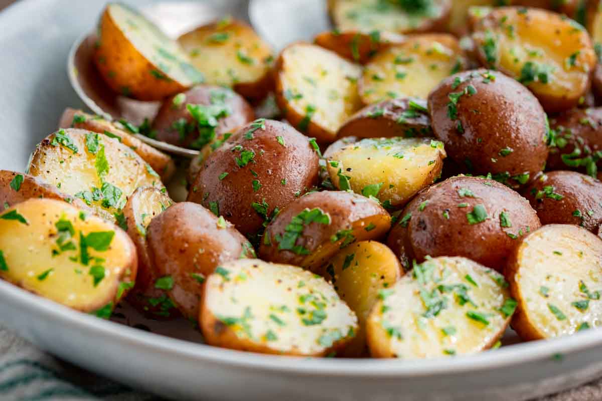 Macro shot of halved baby red potatoes with golden interiors, glistening with butter and scattered with fresh chopped parsley in a white bowl.