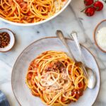 Overhead view of a serving of pasta all'amatriciana in a rustic ceramic bowl, topped with grated pecorino romano, alongside a larger pan of pasta, cherry tomatoes on the vine, red pepper flakes, and a small bowl of grated cheese on a marble surface.