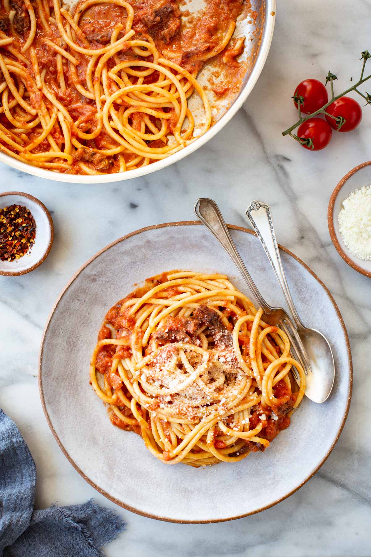 Overhead view of a serving of pasta all'amatriciana in a rustic ceramic bowl, topped with grated pecorino romano, alongside a larger pan of pasta, cherry tomatoes on the vine, red pepper flakes, and a small bowl of grated cheese on a marble surface.
