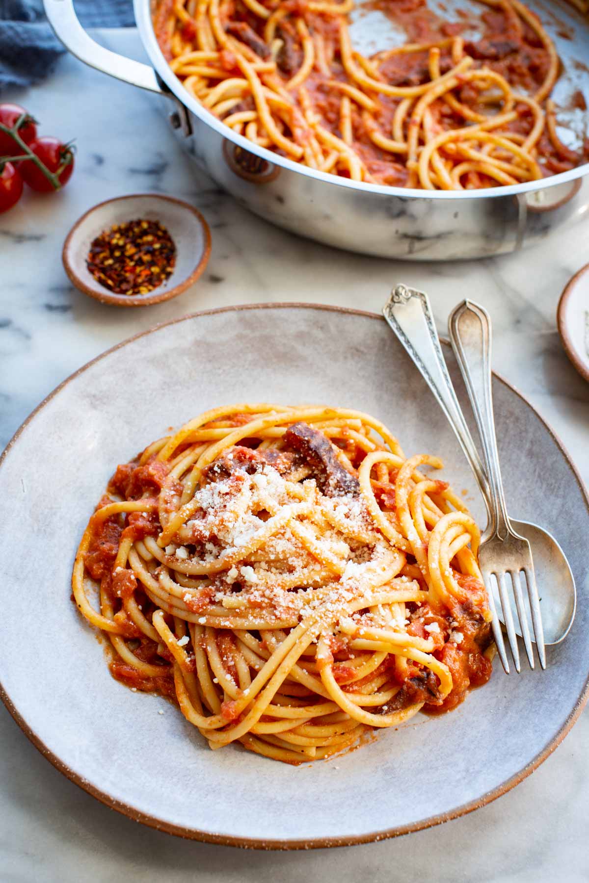 Three-quarter view of a bowl of spaghetti all'amatriciana topped with grated pecorino romano and a visible piece of crisped guanciale, with a stainless steel pan of pasta, red pepper flakes, and cherry tomatoes visible in the background on a marble surface.