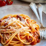 Close-up of a bowl of spaghetti all'amatriciana generously dusted with grated pecorino romano, with a piece of browned guanciale visible among the noodles and a blurred pan of pasta and cherry tomatoes in the background.