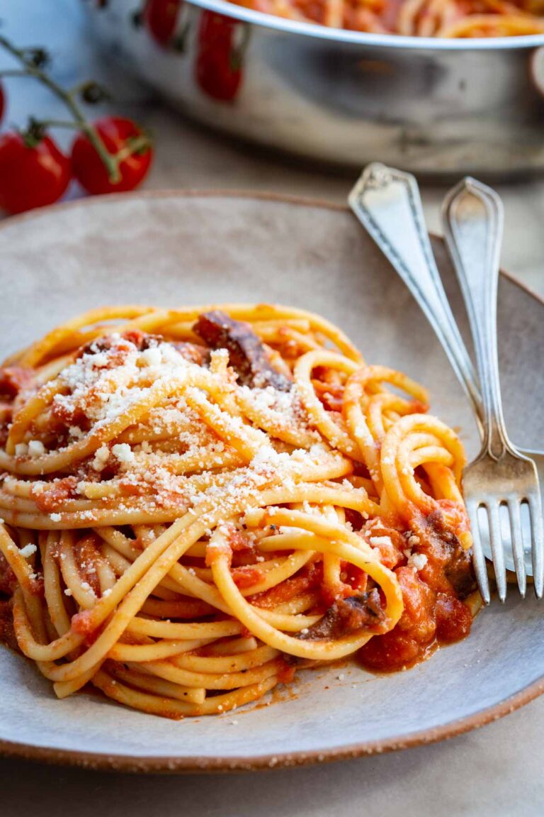 Close-up of a bowl of spaghetti all'amatriciana generously dusted with grated pecorino romano, with a piece of browned guanciale visible among the noodles and a blurred pan of pasta and cherry tomatoes in the background.
