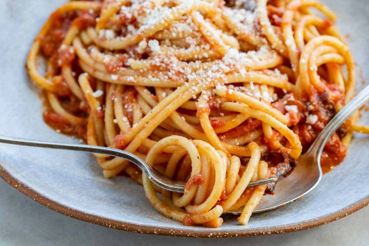 Extreme close-up of spaghetti all'amatriciana being twirled on a fork, showing the thick, glossy tomato sauce, flecks of black pepper, grated pecorino romano, and browned guanciale on a ceramic plate.