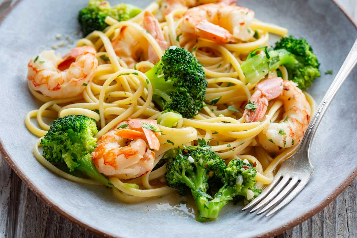 Close-up of shrimp and broccoli pasta on a gray plate, showing plump seasoned shrimp, vivid green broccoli florets, and linguine tossed in a light garlic butter sauce with chopped parsley.