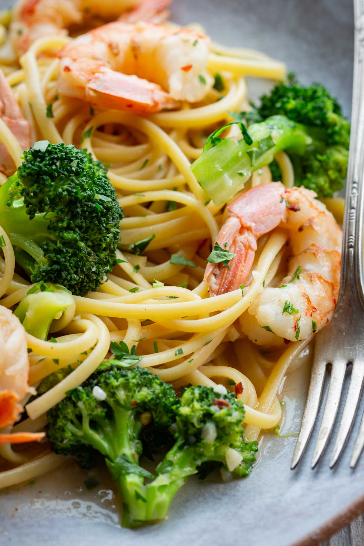 Extreme close-up of shrimp and broccoli linguine on a gray plate, highlighting the glossy garlic butter sauce coating the pasta, tender broccoli florets, and pink shrimp garnished with fresh parsley.