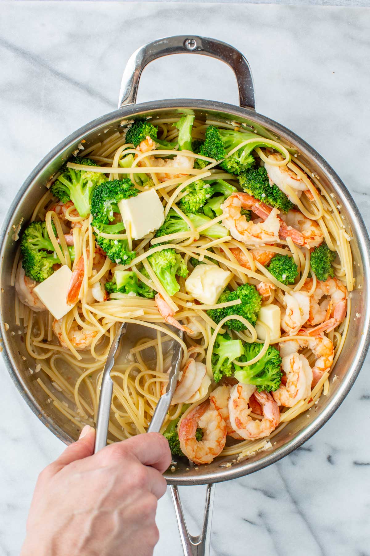 A hand using tongs to toss shrimp and broccoli pasta in a large stainless steel skillet, with cubes of butter melting into the linguine on a marble surface.