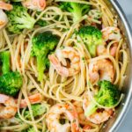 Close-up overhead view of shrimp and broccoli linguine in a stainless steel skillet, with plump pink shrimp, bright green broccoli florets, and flecks of fresh parsley scattered throughout.