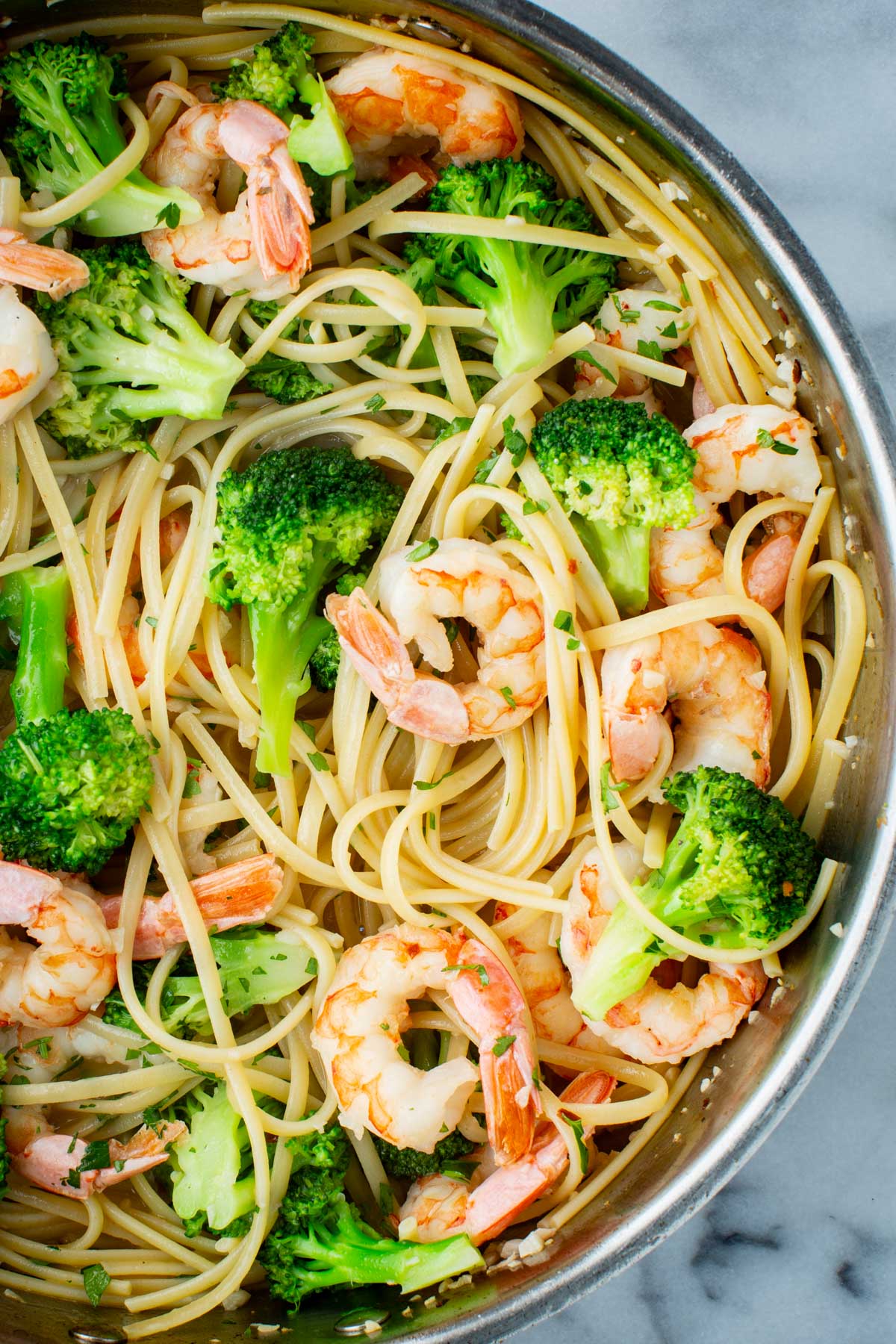 Close-up overhead view of shrimp and broccoli linguine in a stainless steel skillet, with plump pink shrimp, bright green broccoli florets, and flecks of fresh parsley scattered throughout.
