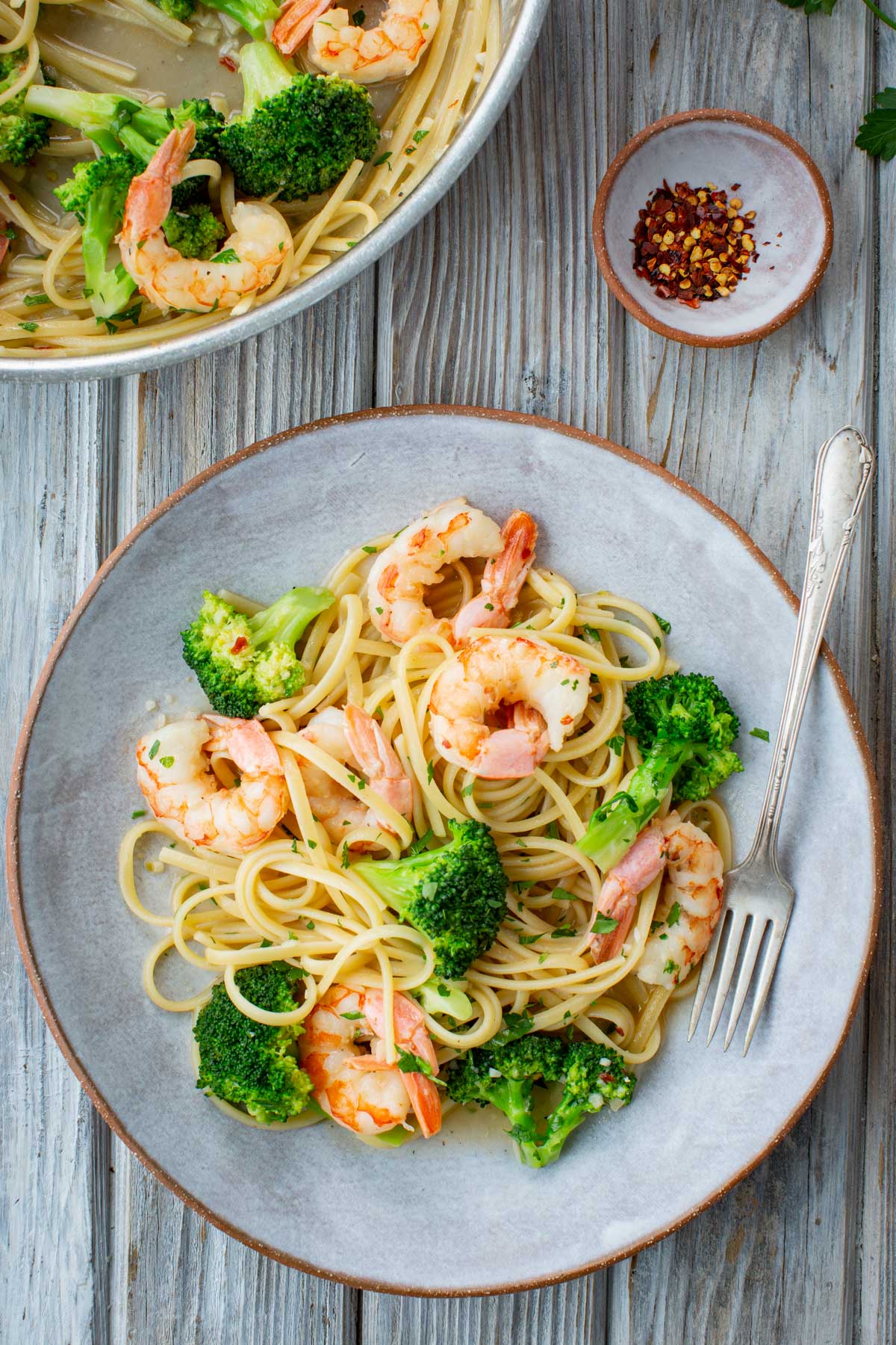 Overhead view of a plated serving of shrimp and broccoli linguine on a rustic gray plate with a silver fork, alongside a small clay bowl of red pepper flakes and the skillet in the background.