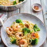 A plate of shrimp and broccoli pasta on a weathered wood surface, with a silver fork, a small dish of red pepper flakes, a linen napkin, and fresh parsley sprigs in the background.