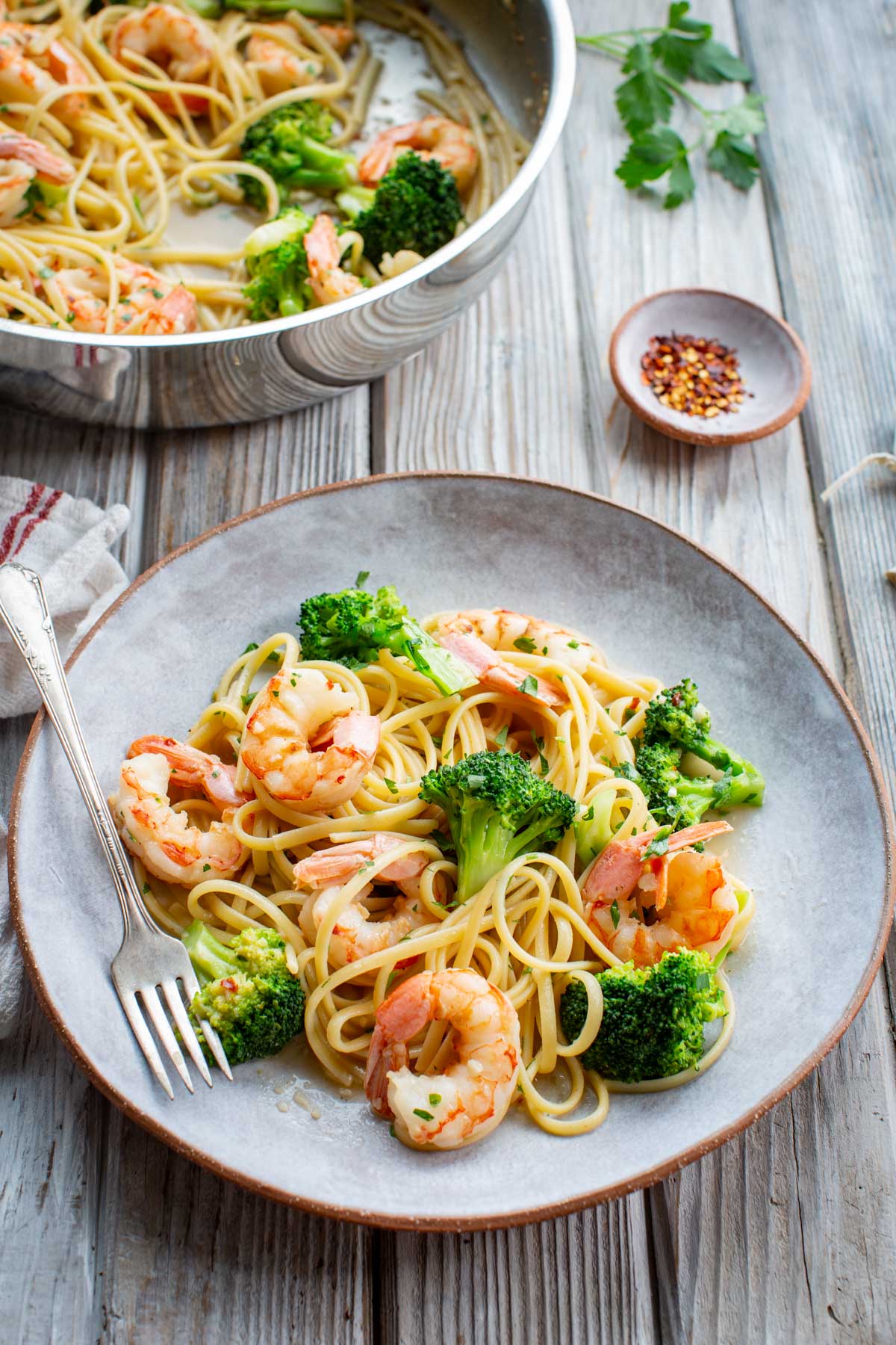 A plate of shrimp and broccoli pasta on a weathered wood surface, with a silver fork, a small dish of red pepper flakes, a linen napkin, and fresh parsley sprigs in the background.