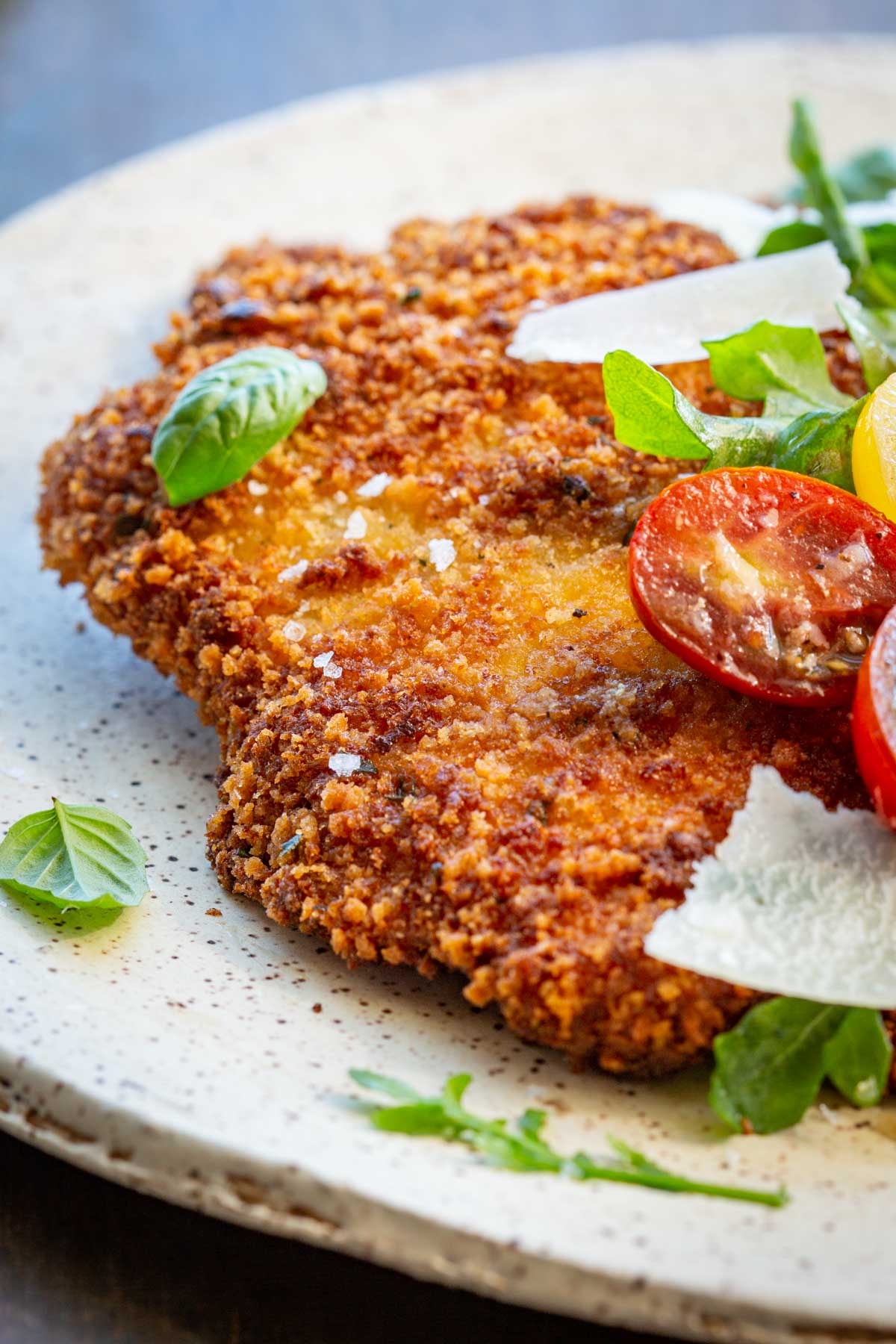 Extreme close-up of a crispy veal Milanese cutlet on a ceramic plate, showing the crunchy golden crust with flaky sea salt, garnished with fresh basil leaves, halved cherry tomatoes, and shaved parmesan.