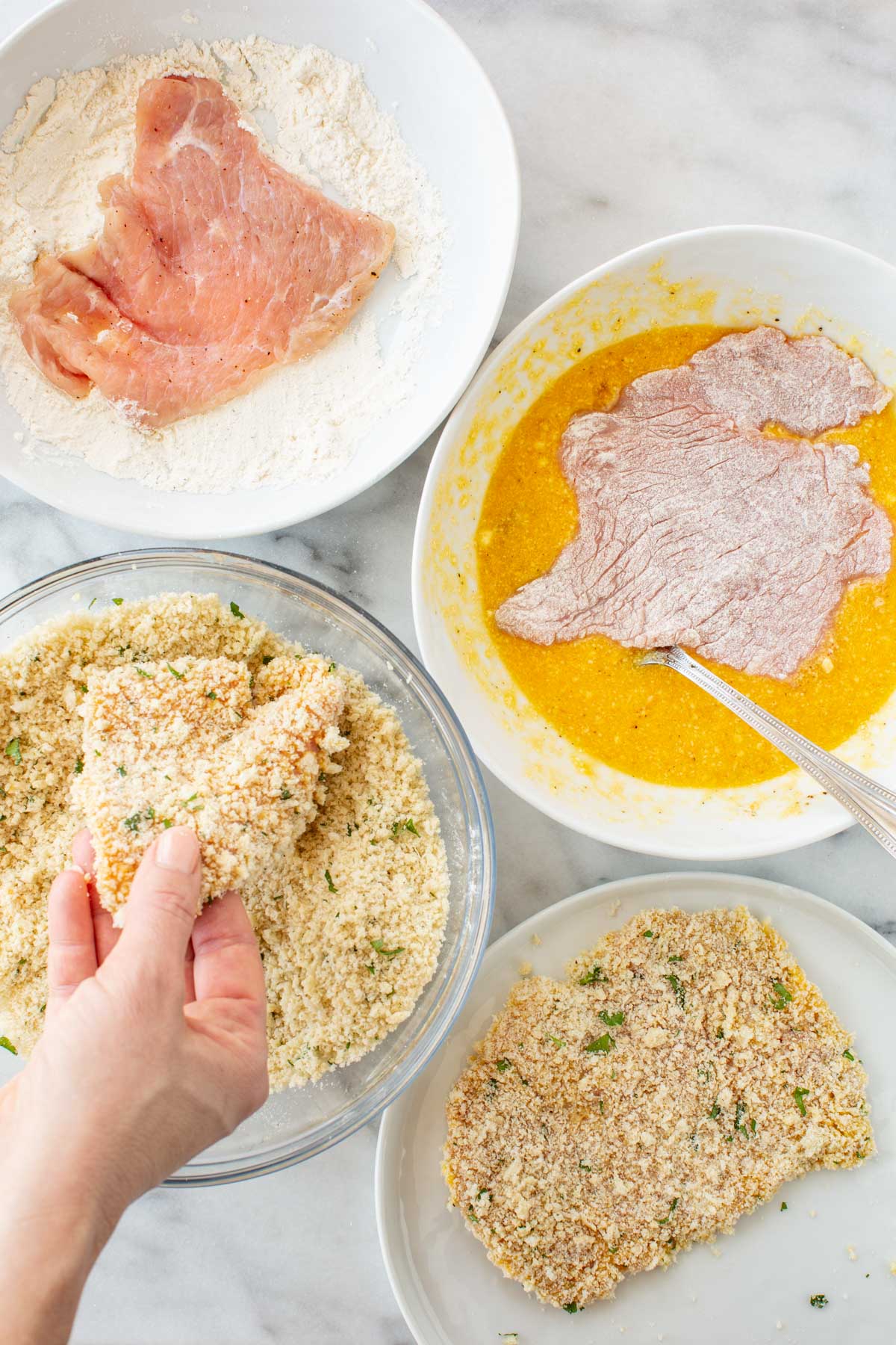 Overhead view of a three-bowl breading station: seasoned flour with veal cutlets (top left), beaten egg wash with a cutlet soaking (top right), and a bowl of herbed breadcrumb mixture (left), with a finished breaded cutlet resting on a plate (bottom right).