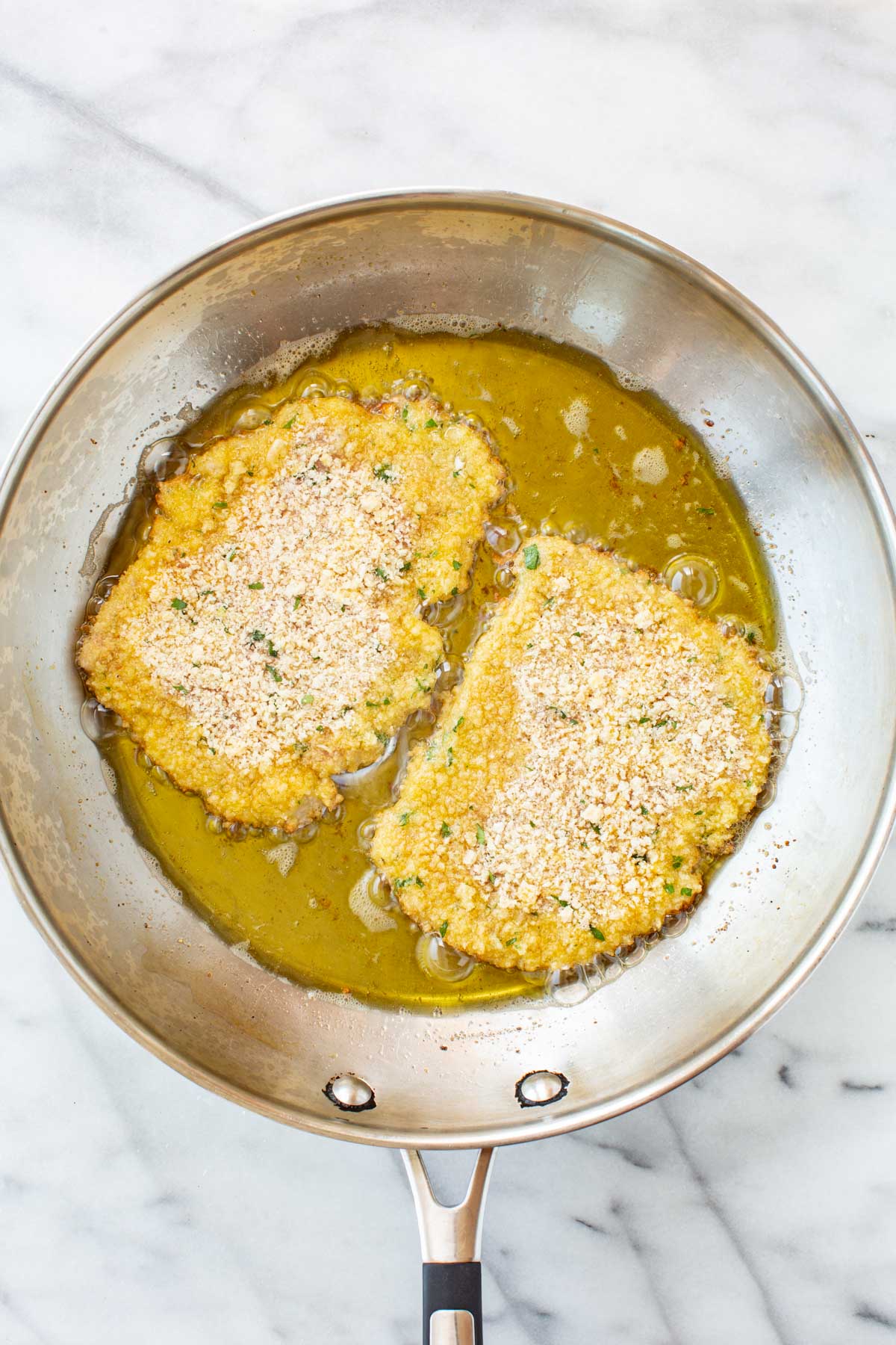 Two breaded veal cutlets frying in a pool of olive oil in a stainless steel skillet, with the breadcrumbs still pale and untoasted on the top side.