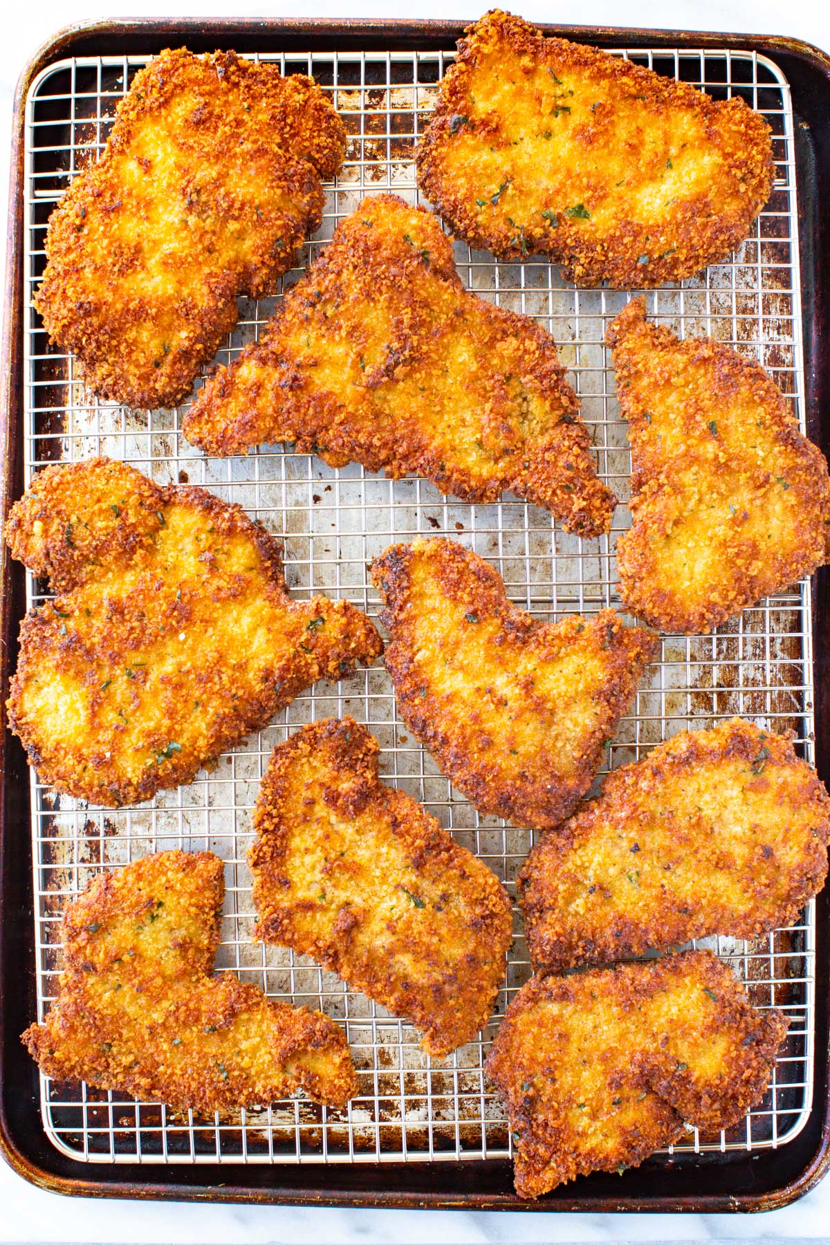 Overhead shot of eight golden-brown, crispy pan-fried veal cutlets draining on a wire rack set over a baking sheet.