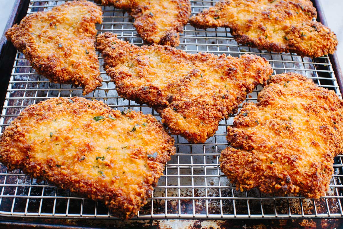 Close-up wide-angle view of multiple crispy veal Milanese cutlets resting on a wire rack, showing their deeply golden, textured breadcrumb crust.
