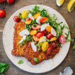Overhead shot of a single veal Milanese cutlet on a rustic ceramic plate, topped with a fresh arugula, cherry tomato, and shaved parmesan salad, surrounded by loose cherry tomatoes, fresh basil leaves, and lemon wedges on a wooden surface.