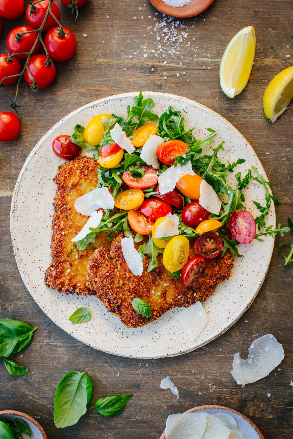 Overhead shot of a single veal Milanese cutlet on a rustic ceramic plate, topped with a fresh arugula, cherry tomato, and shaved parmesan salad, surrounded by loose cherry tomatoes, fresh basil leaves, and lemon wedges on a wooden surface.