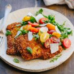 Angled view of a plated veal Milanese cutlet topped with arugula, colorful halved cherry tomatoes, and shaved parmesan, served on a speckled ceramic plate with a vintage fork, on a wooden table.