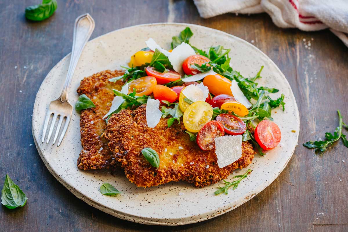 Angled view of a plated veal Milanese cutlet topped with arugula, colorful halved cherry tomatoes, and shaved parmesan, served on a speckled ceramic plate with a vintage fork, on a wooden table.